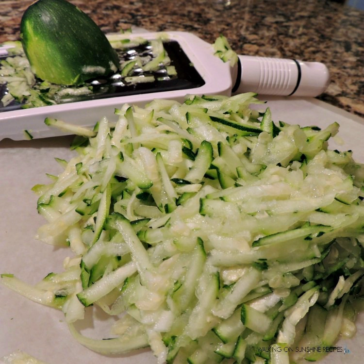 Grated zucchini on white plastic cutting board with grater in background. 
