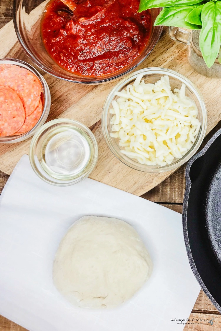 Dough base with bowls of marinara sauce, pepperoni and mozzarella cheese. 