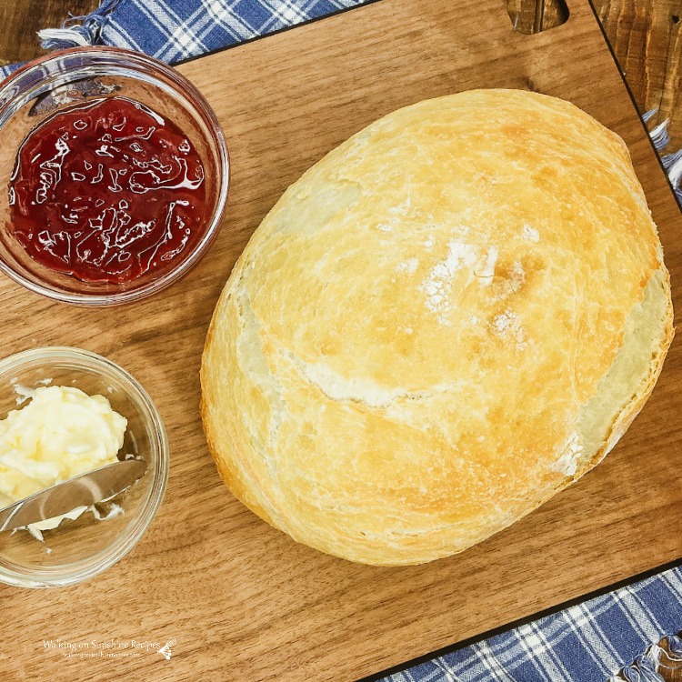 Rustic loaf of No Knead Bread on Cutting Board with jam and butter in small bowls. 