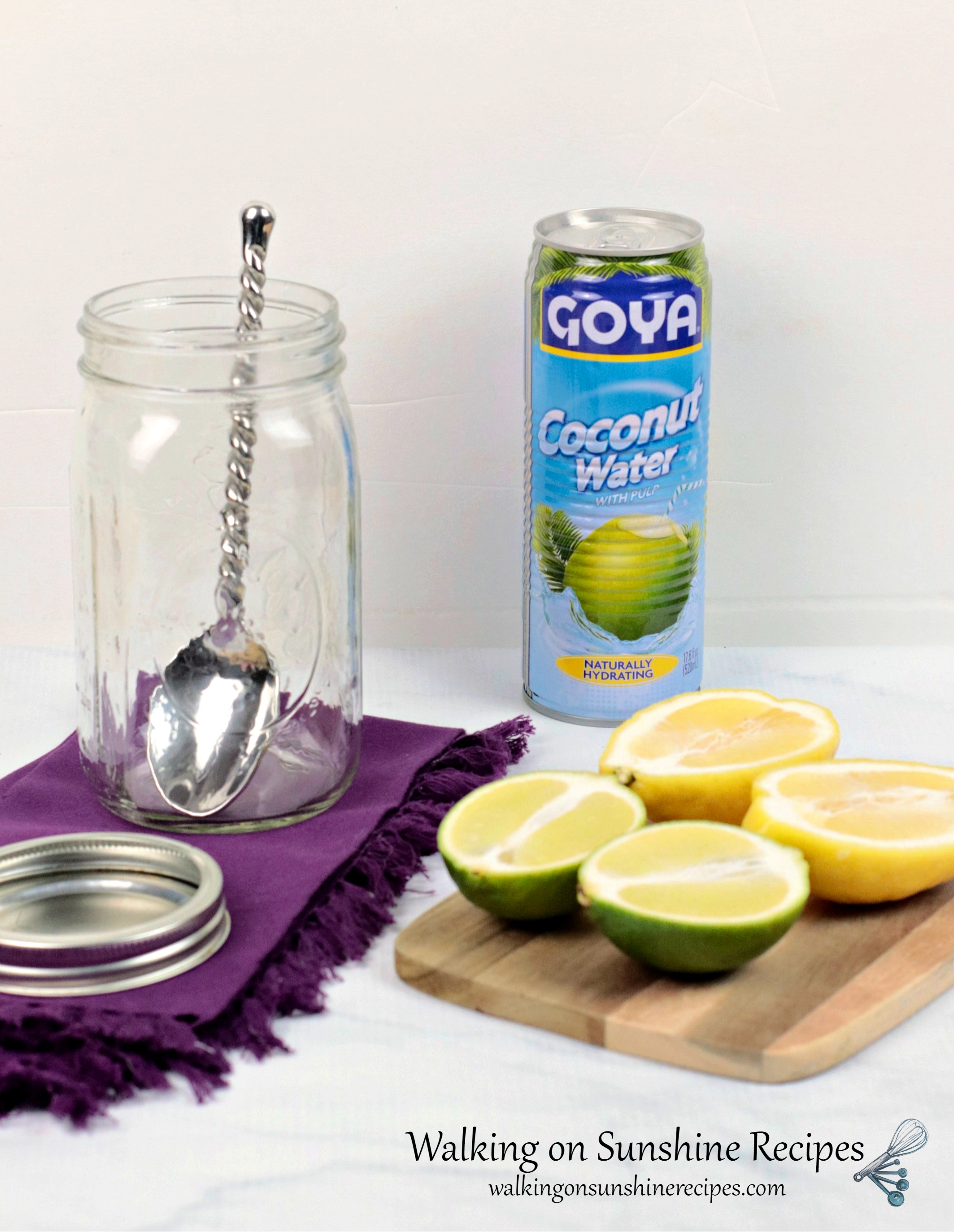Lemon lime water ingredients and tools. Can of Goya coconut water, lemon cut in half, lime cut in half displayed on wood cutting board. Empty mason jar with large metal spoon inside on top of a purple cloth napkin. 