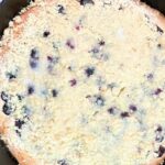 A golden brown cornbread topped with blueberries and a crumb topping in a cast iron skillet.