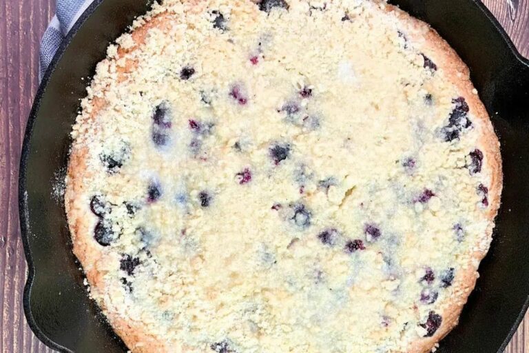 A golden brown cornbread topped with blueberries and a crumb topping in a cast iron skillet.
