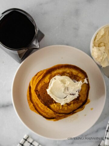 Top view of pumpkin pancakes with butter on top. A pitcher of syrup to the left and ramekin of butter to the right of the plate of pancakes.
