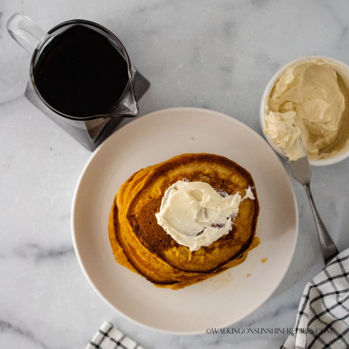 Top view of pumpkin pancakes with butter on top. A pitcher of syrup to the left and ramekin of butter to the right of the plate of pancakes.