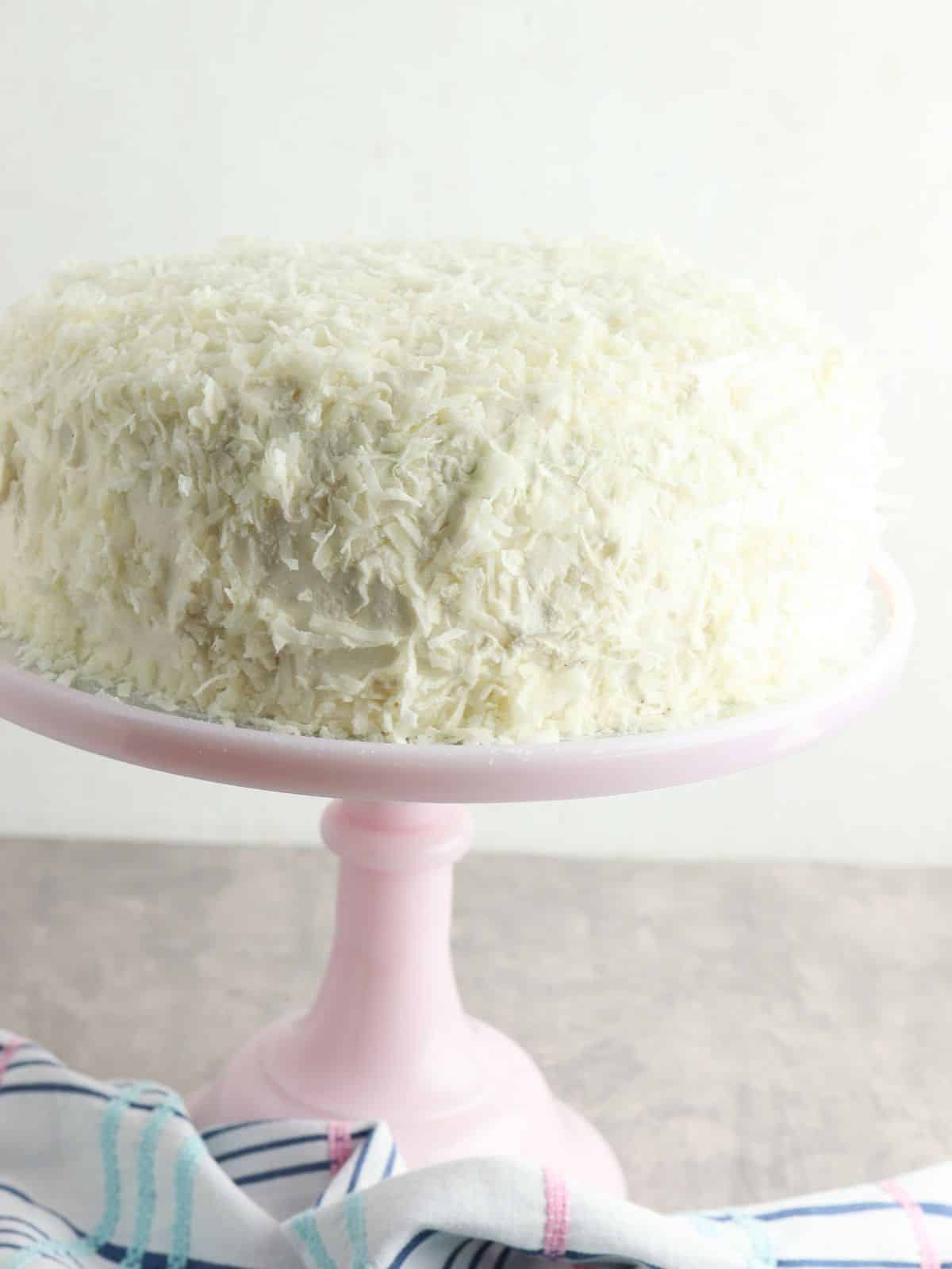 cake with cream cheese frosting and shredded coconut on pink cake stand.