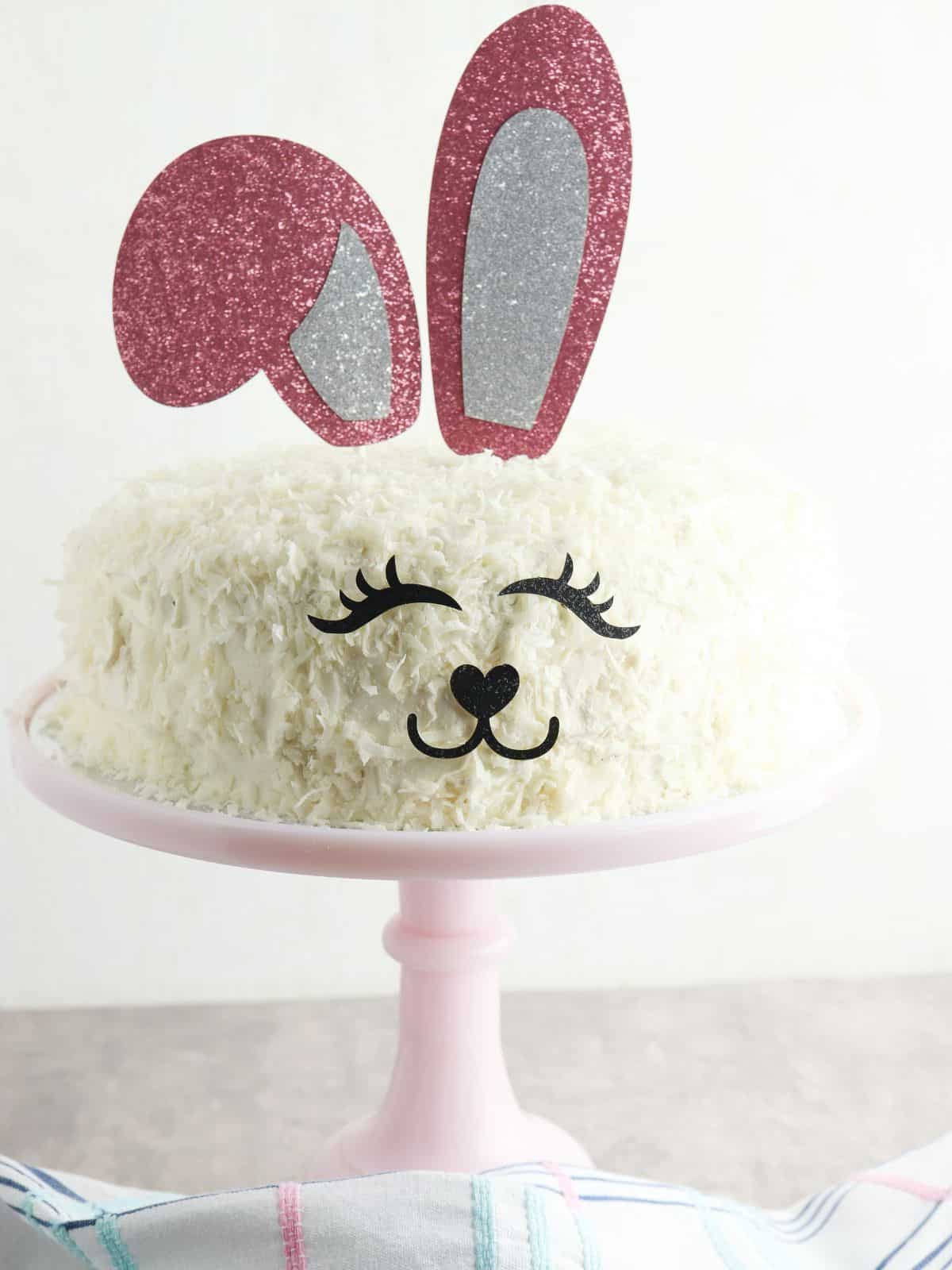 round cake with bunny ears, eyes and nose on pink cake stand.