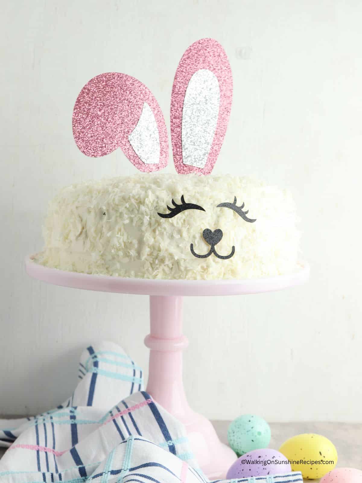Easter bunny cake on pink cake stand with colored eggs on table top.