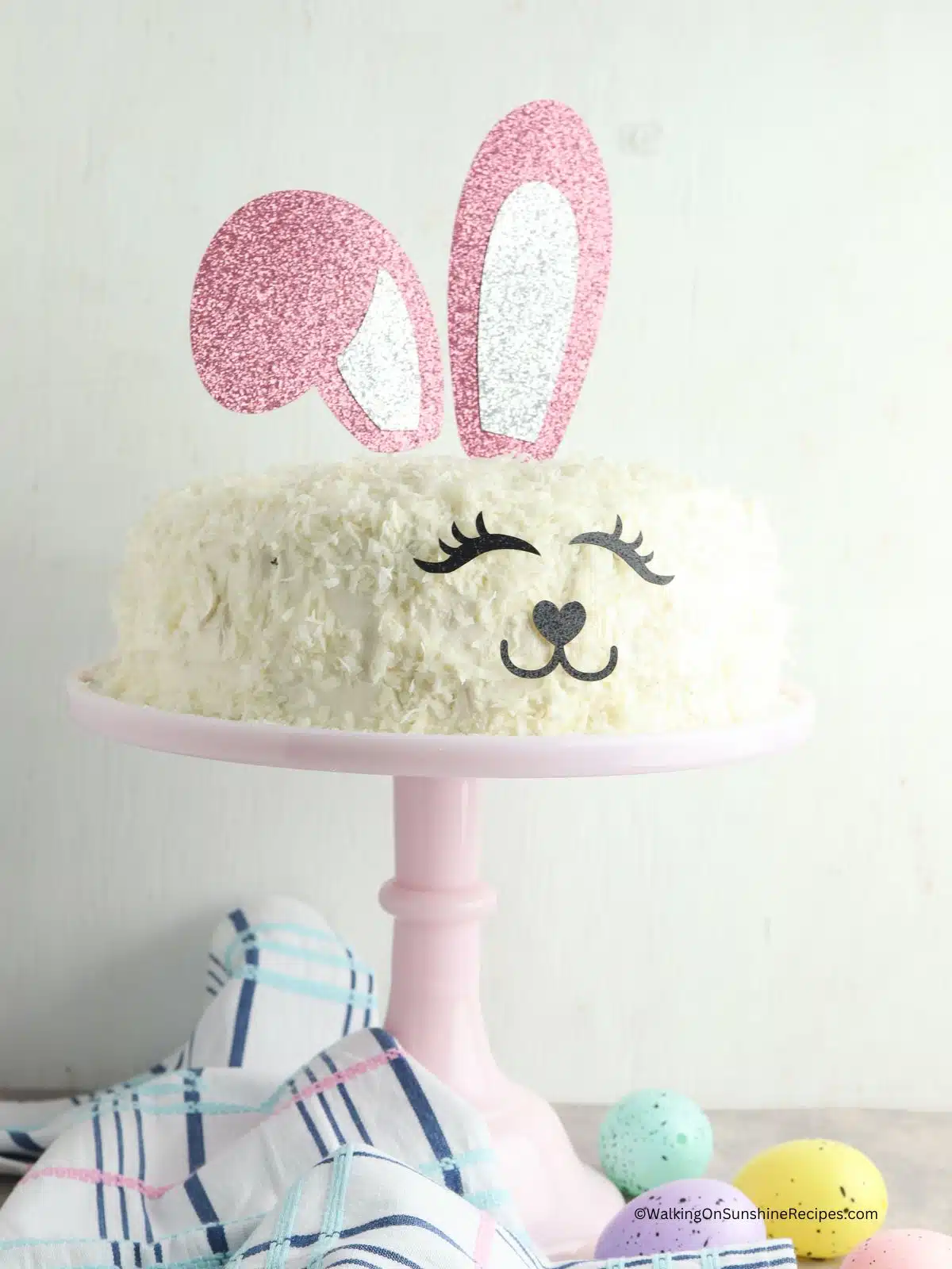 Easter bunny cake on pink cake stand with colored eggs on table top.