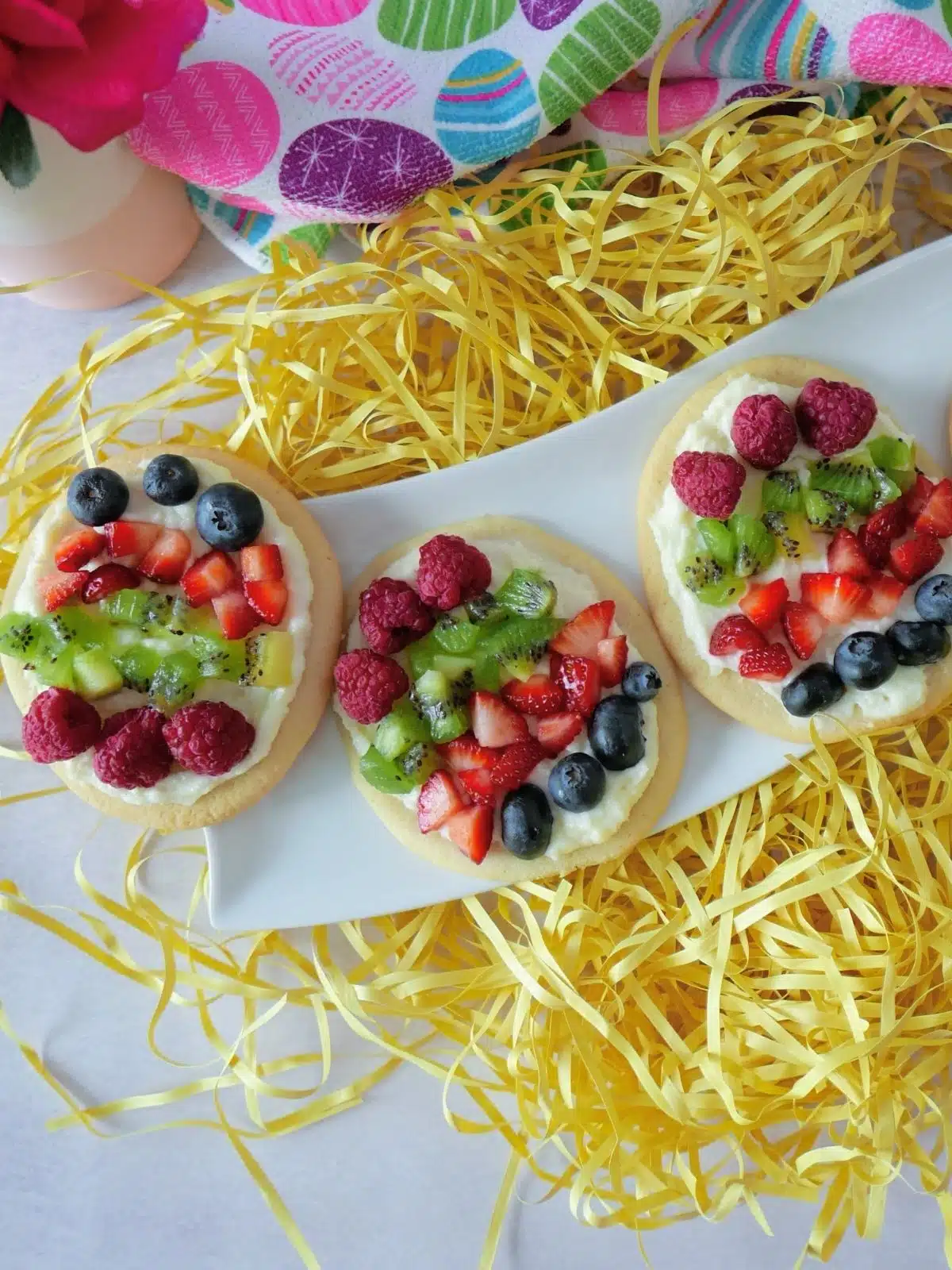 fruit and cream cheese on sugar cookies shaped like eggs on white tray and yellow grass.