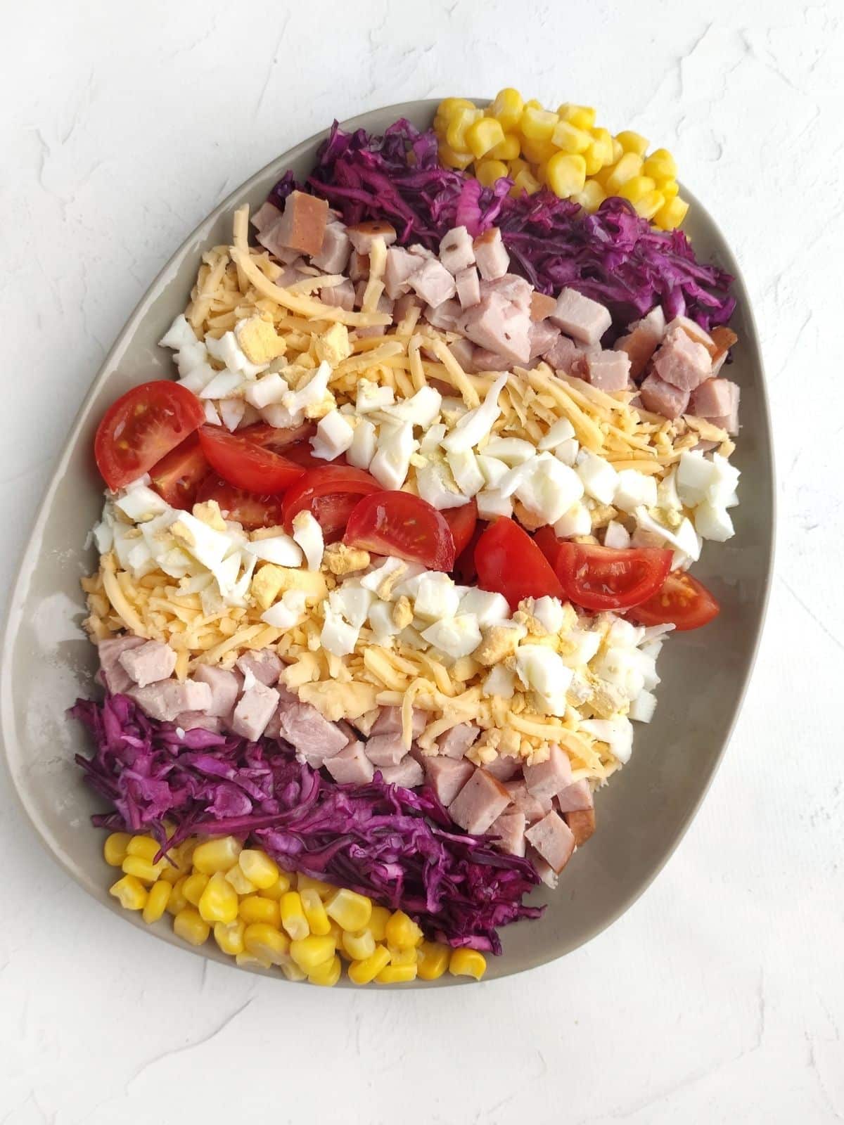 Salad on egg shaped plate with corn, red cabbage, tomato, cheese and ham.
