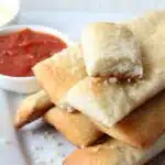 pizza dough breadsticks on cutting board with pizza sauce in white bowl.
