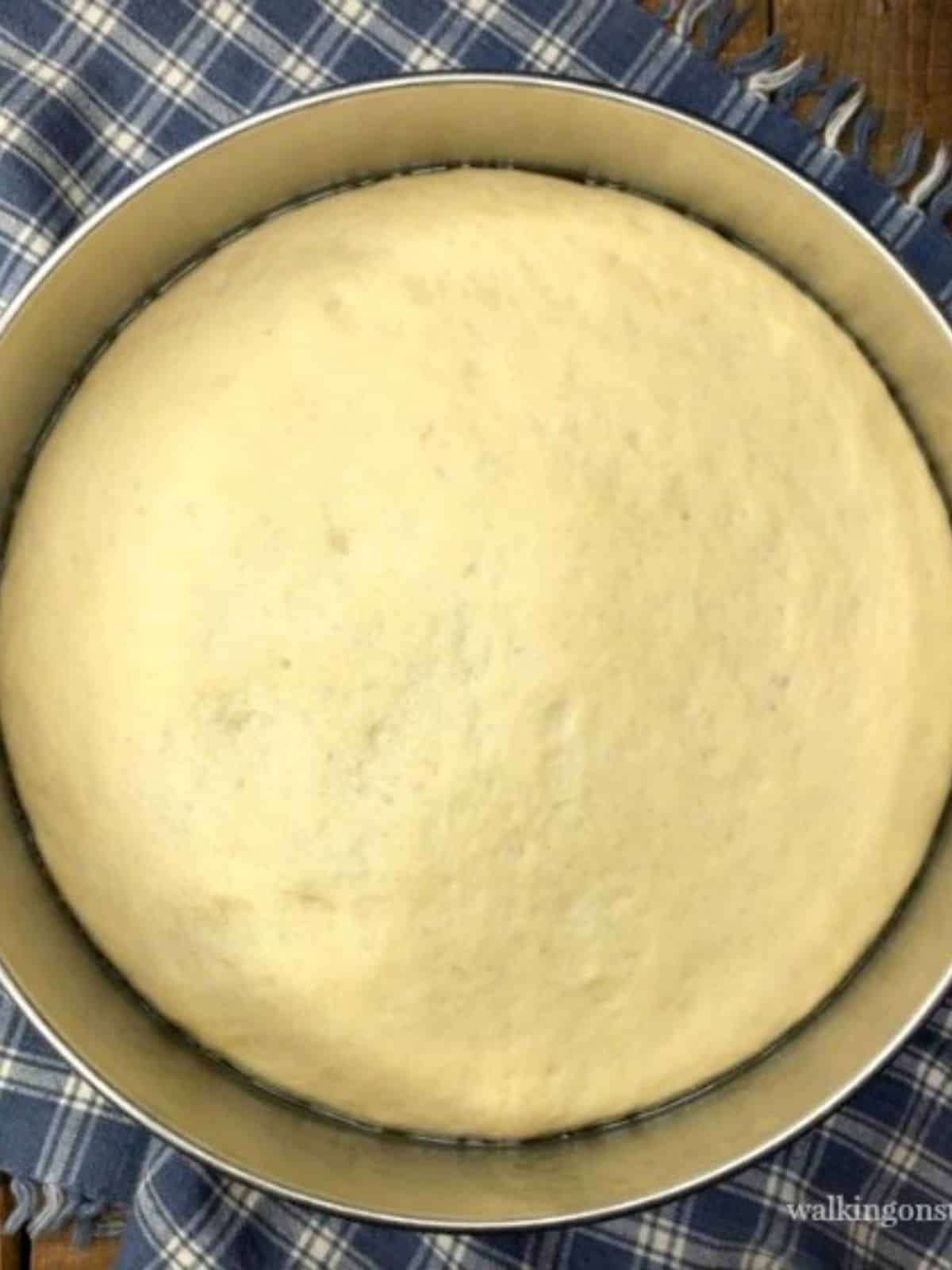 risen pizza dough in metal bowl on blue plaid napkin.
