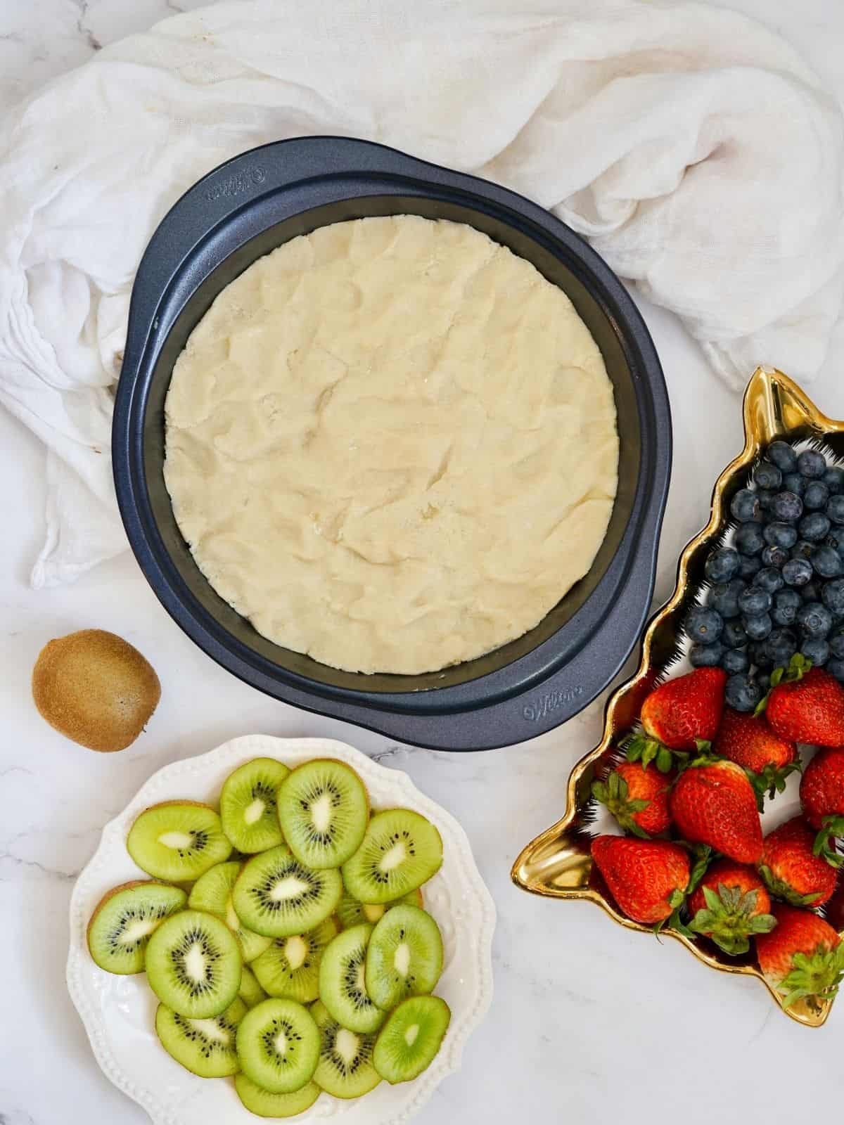 sugar cookie dough spread out in cake pan with fruit in bowl and plate nearby.