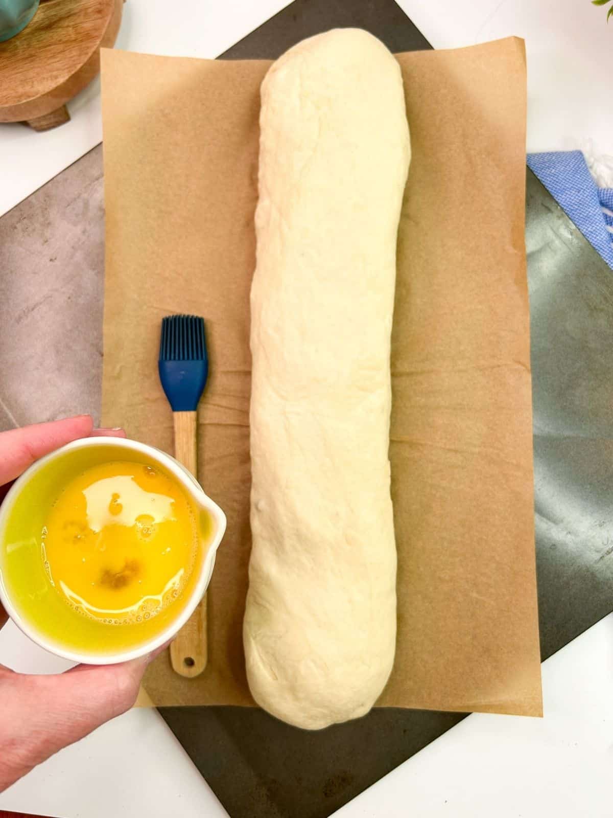 beaten egg in small dish held over rolled up sausage roll on parchment paper.
