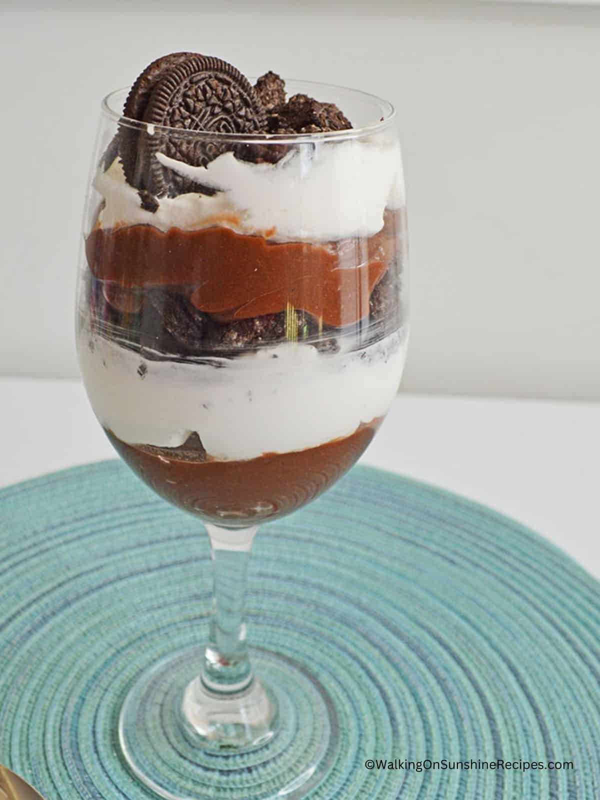 Oreo pudding parfait in clear glass on aqua placemat.