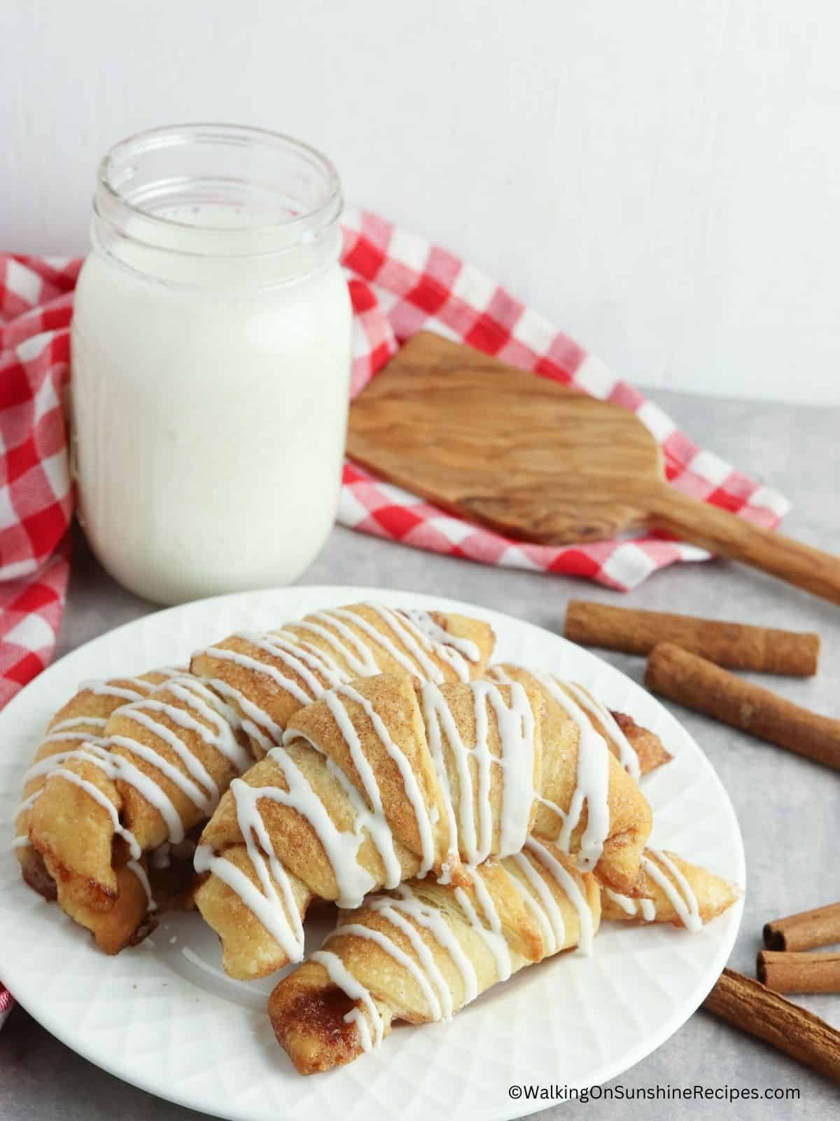 cinnamon crescent rolls with cream cheese frosting drizzle on white plate with cinnamon sticks and glass of milk.