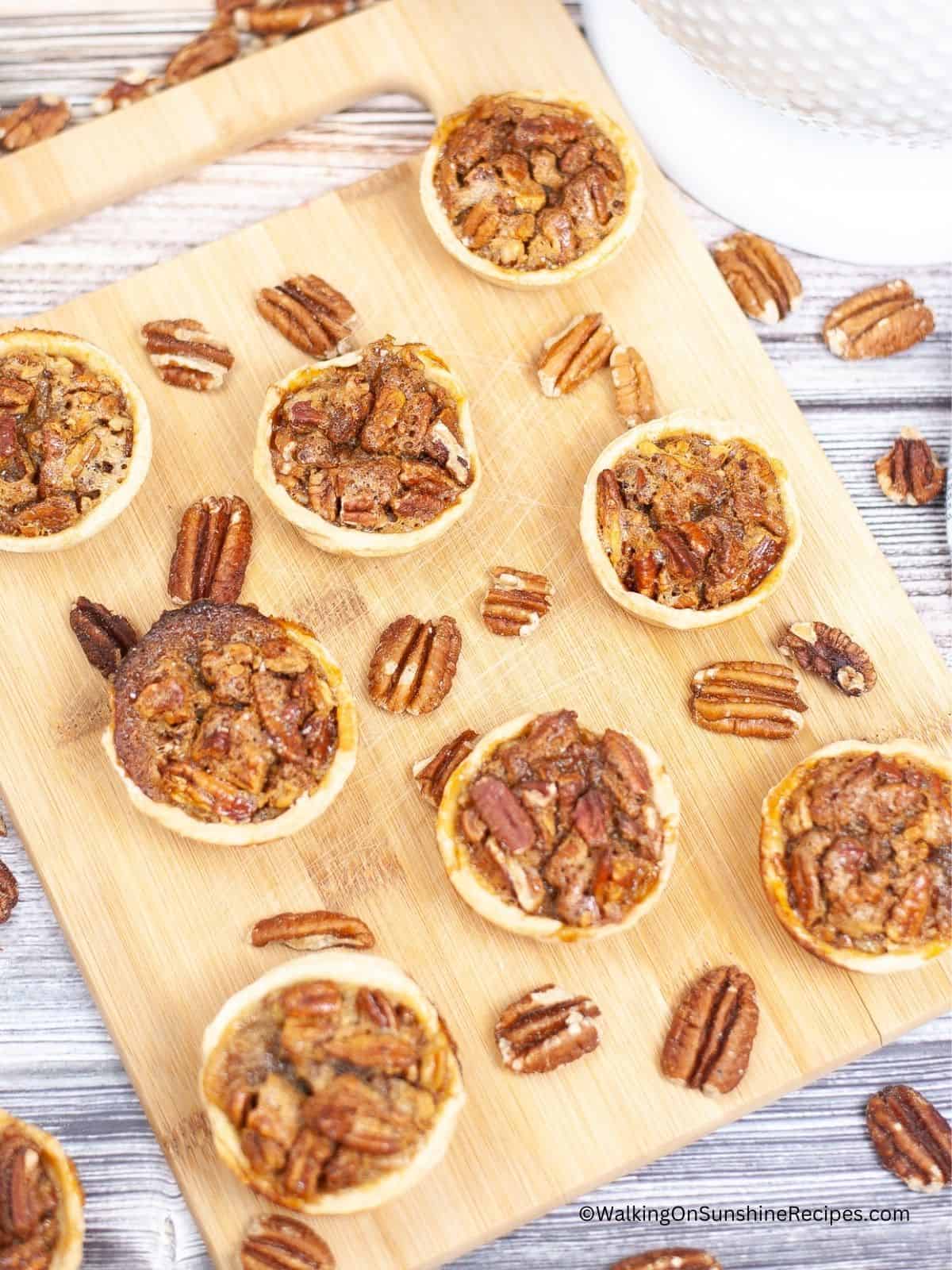 mini pecan pies on cutting board with lose pecans surrounding.