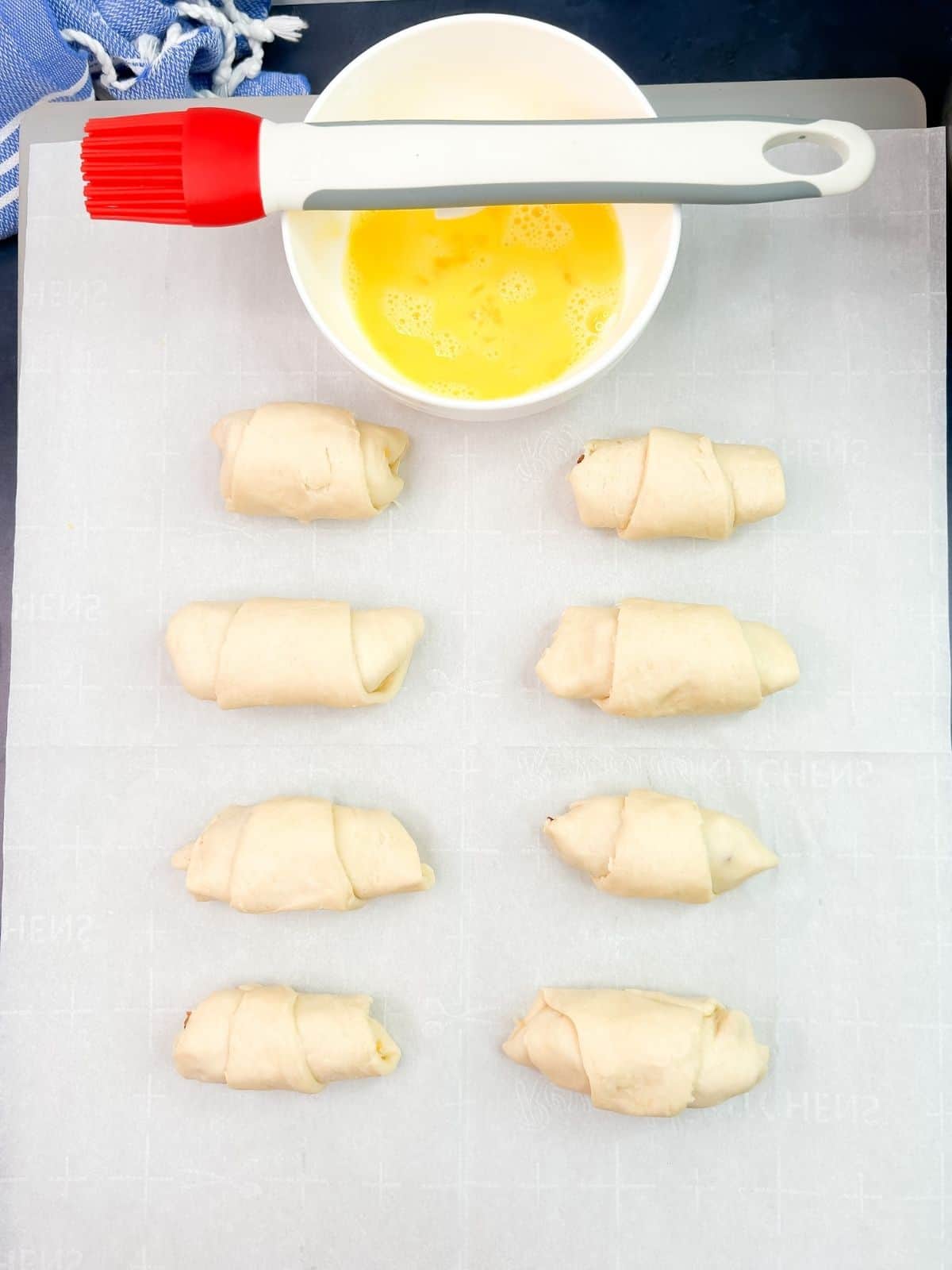unbaked crescent rolls on baking tray with bowl of egg and silicone brush.