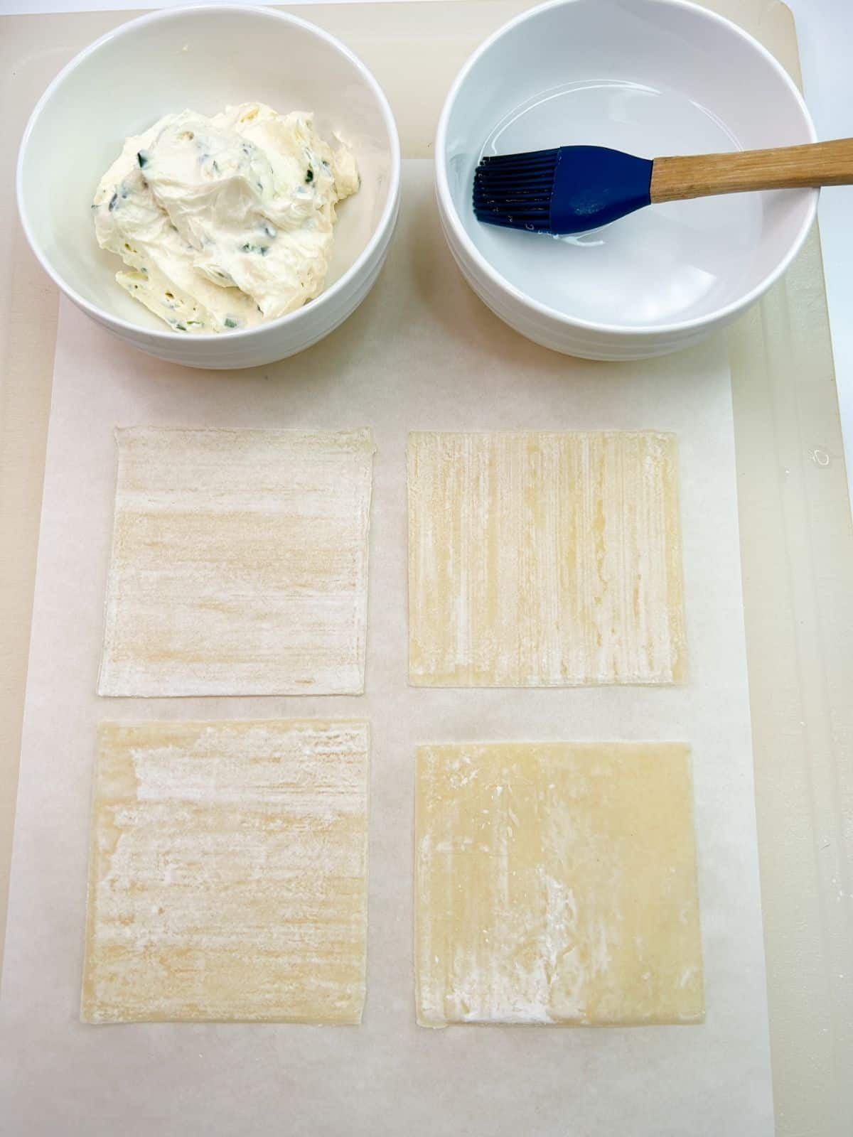 4 won ton wrappers on parchment paper with cream cheese mixture in bowl and water in bowl.