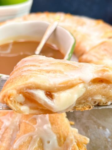 Slice of crescent roll filled with apple pie filling and cream cheese on a spatula with a bowl of caramel the rest of the crescent ring in the backgroung.