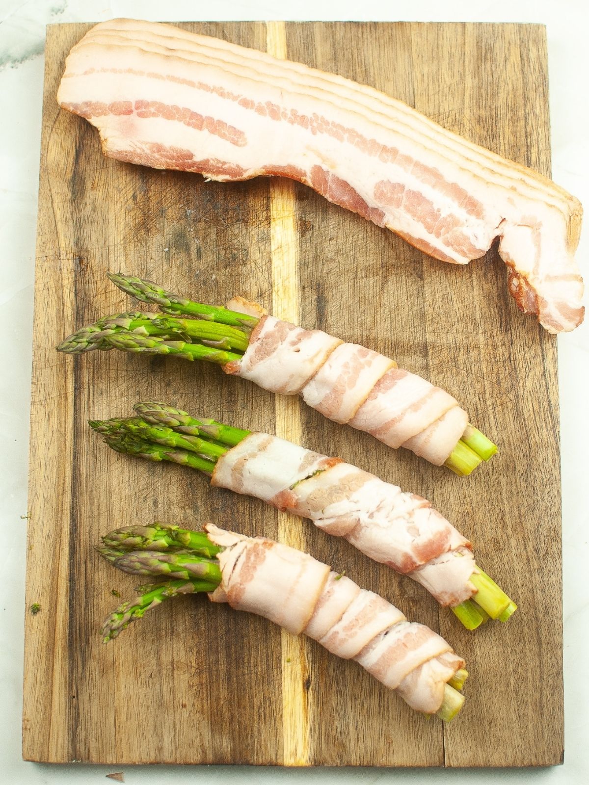 3 bunches of asparagus wrapped in raw bacon on cutting board.