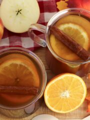 overhead photo of apple cider with apples and oranges in glass mugs.