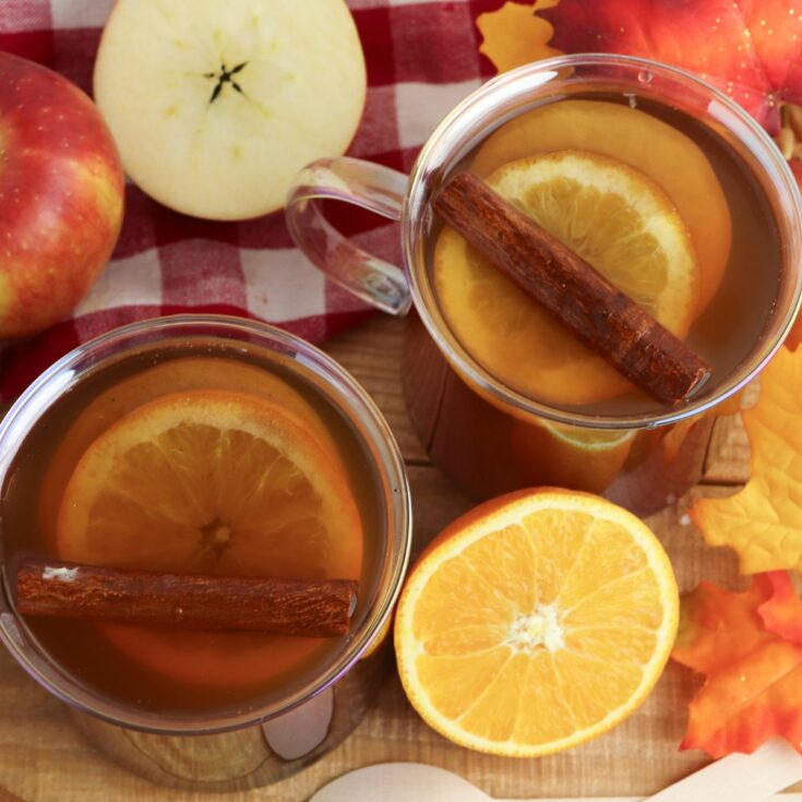 overhead photo of apple cider with apples and oranges in glass mugs.