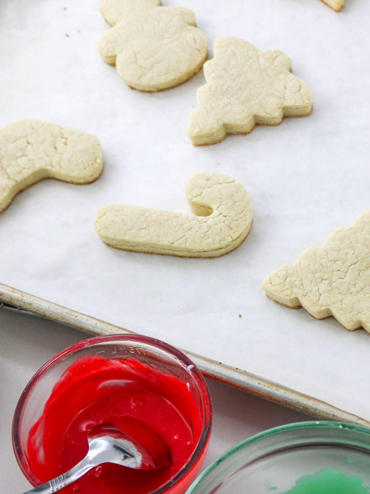 Baked cut out Christmas cookies on tray with bowls of red and green frosting.