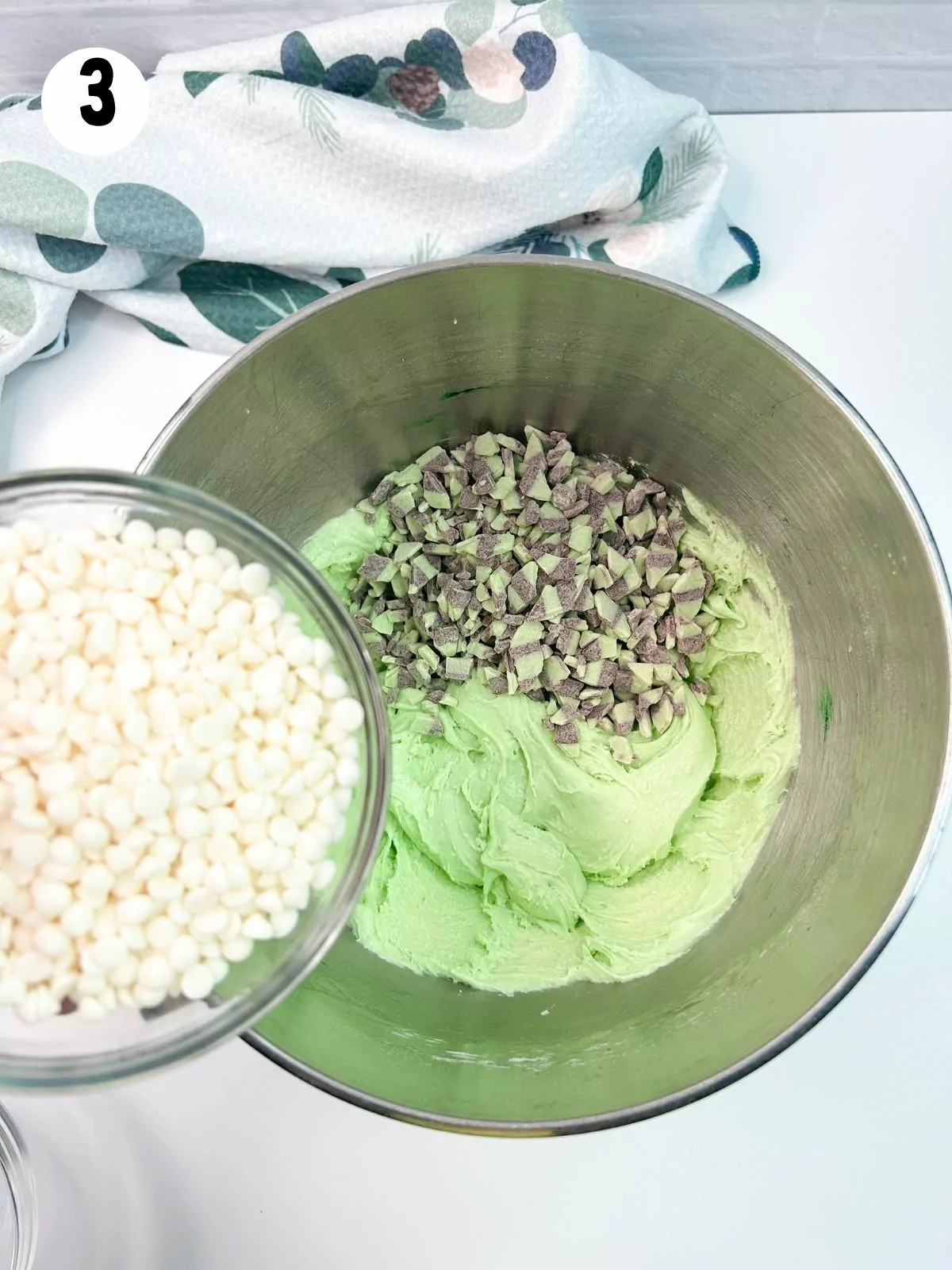 Mini white chocolate chips in bowl being added to green cake mix batter.