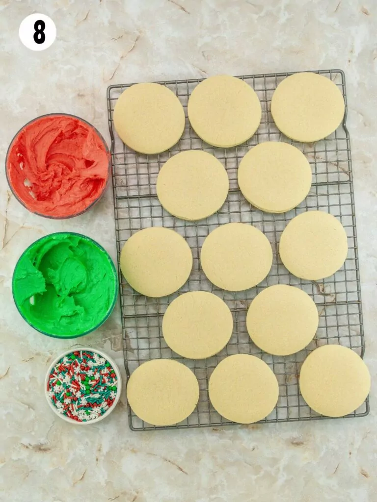 sugar cookies on cooling rack with frosting and sprinkles in small bowls.