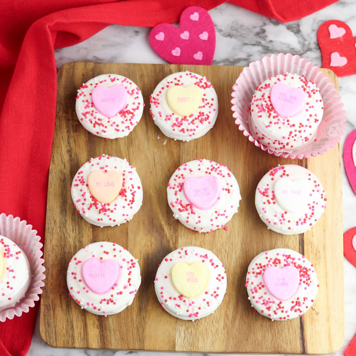 Square photo Oreos dipped in melted chocolate on cutting board with sprinkles and candy hearts on top.