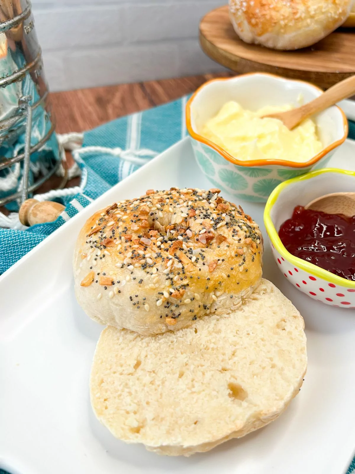 Bagel with everything bagel seasoning sliced open on white dish with butter, jam and cream cheese in small bowls.