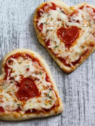 two home-baked pizza in the shape of hearts on a gray cutting board.