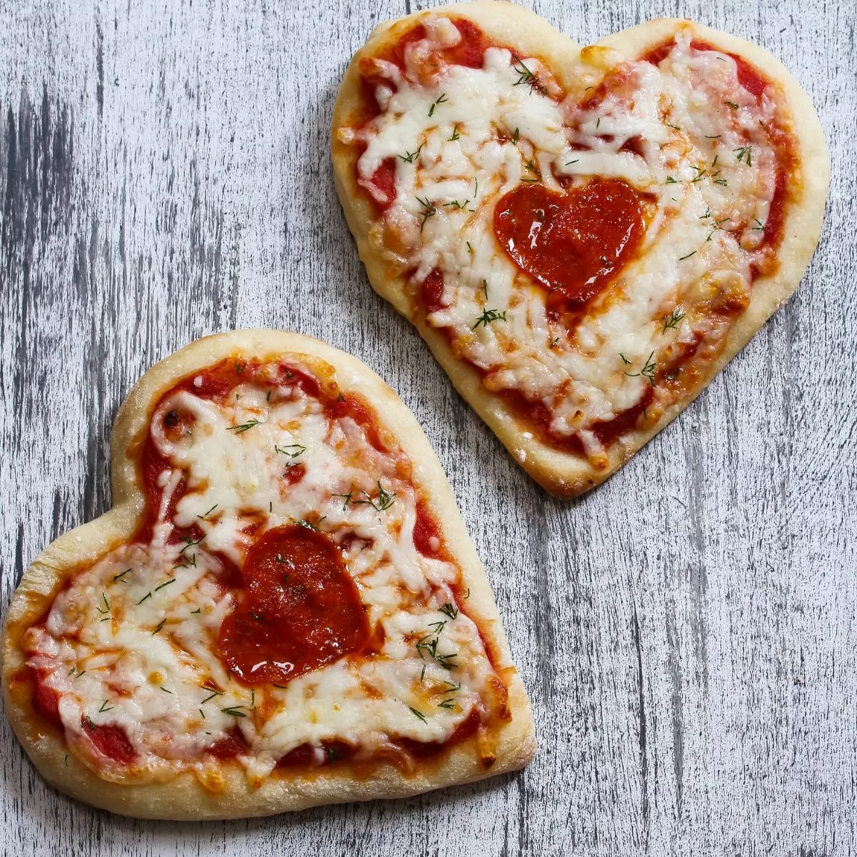 two home-baked pizza in the shape of hearts on a gray cutting board.