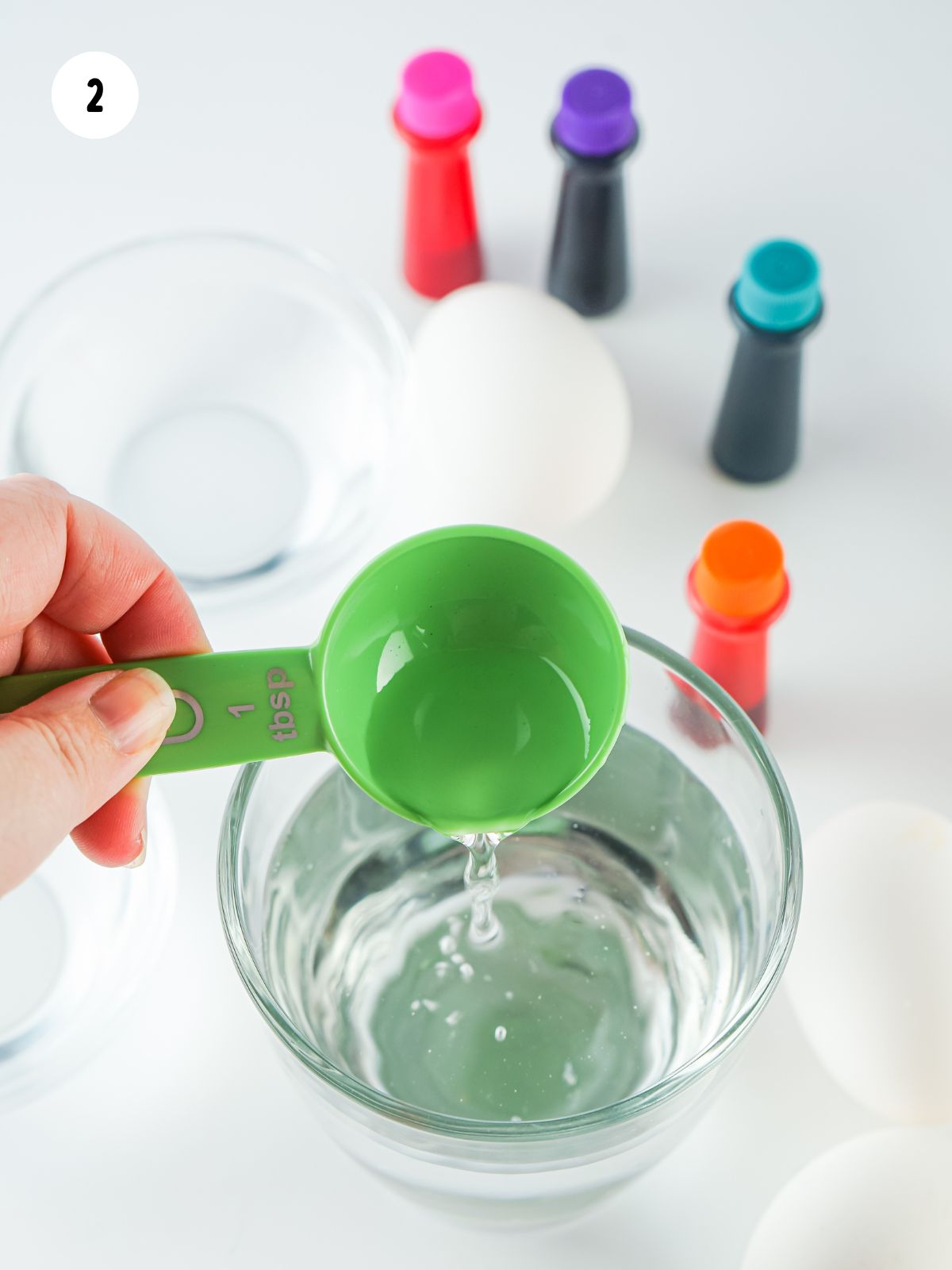 adding vinegar to glass of water with bottles of food coloring in the background.