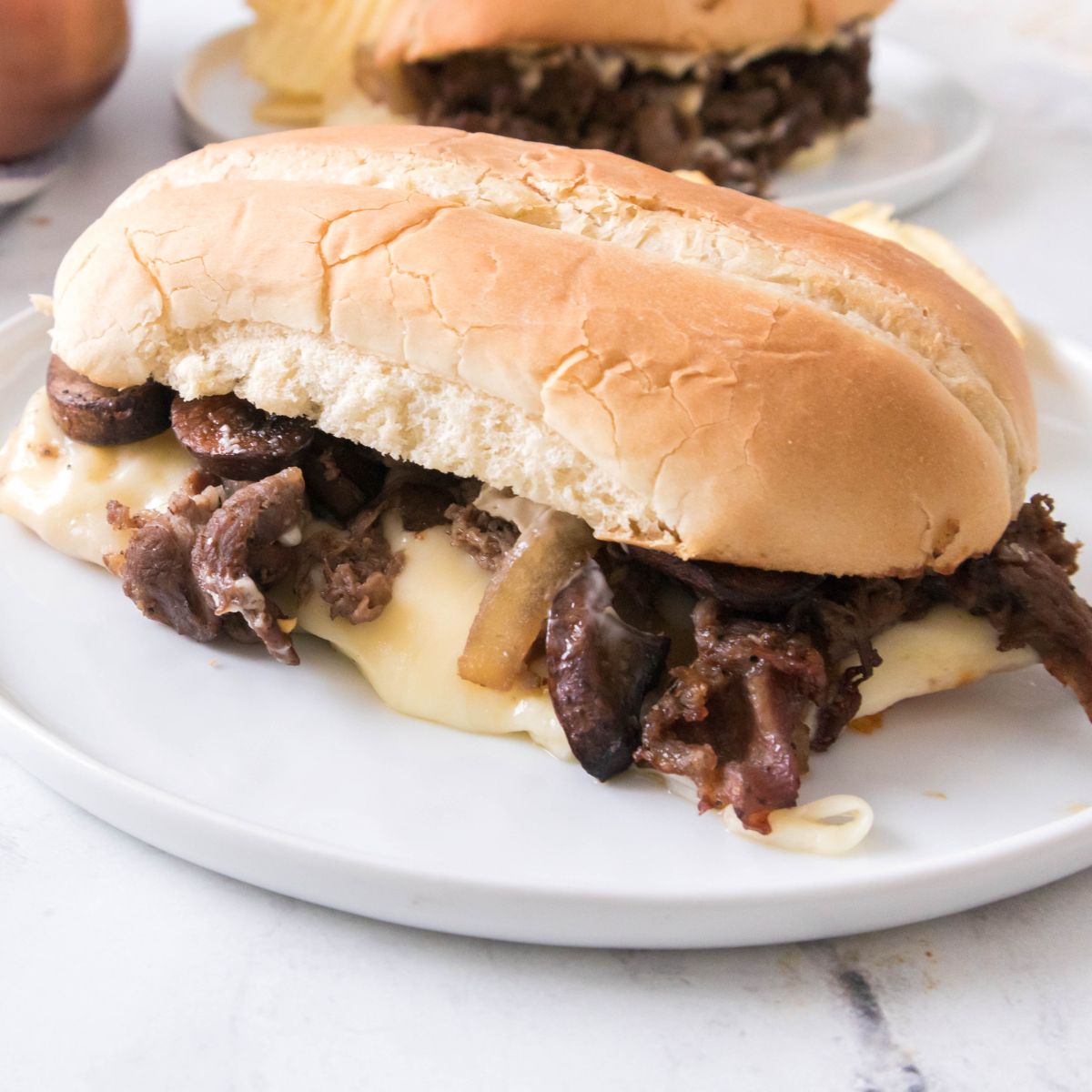 Philly cheesesteak with provolone cheese, mushrooms and onions on hoagie roll on white plate.