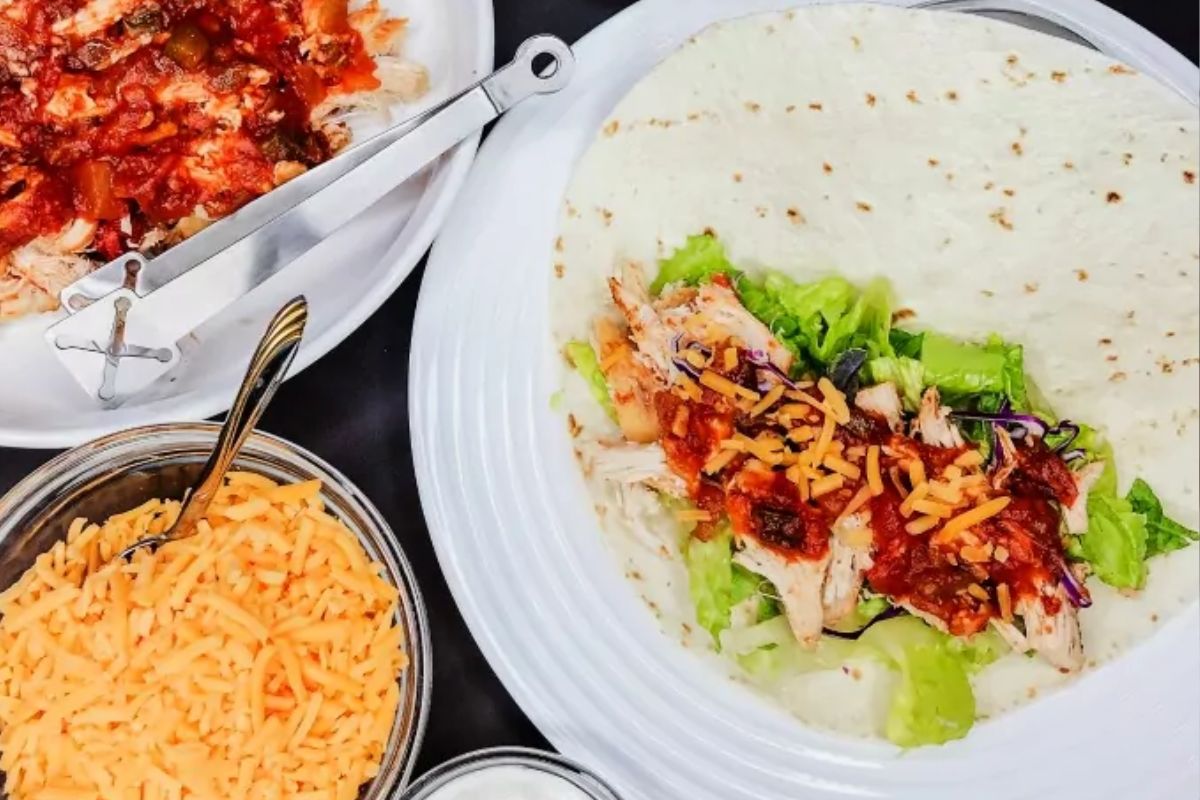 Shredded chicken topped with salsa, cheddar cheese and lettuce in a flour tortilla. A bowl of cheddar cheese to the side.