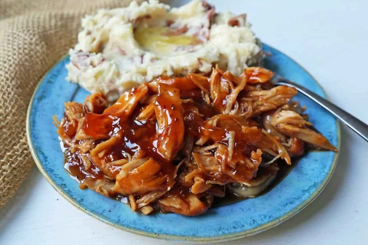 shredded chicken with barbecue sauce on blue plate with smashed potatoes in the back.