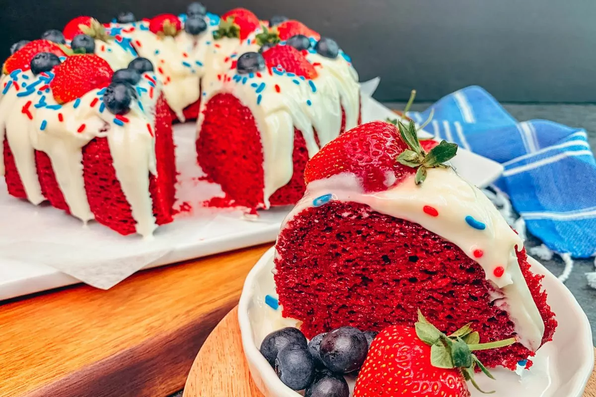 A slice of red, velvet cake decorated with strawberries and blueberries. The whole cake is in the background.