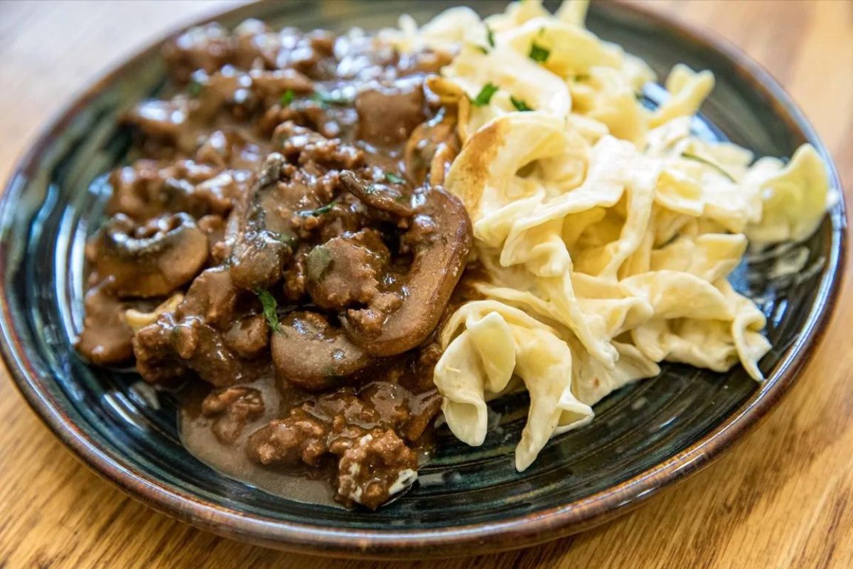 beef, mushrooms with gravy served next to pasta with cream sauce on plate.