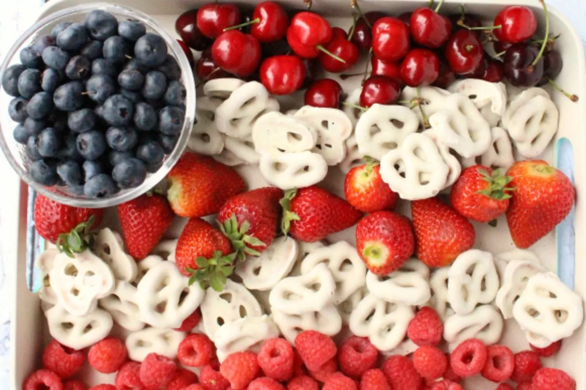 wooden tray with raspberries, strawberries, cherries, white chocolate covered pretzels and blueberries all laid out in the shape of an American flag.