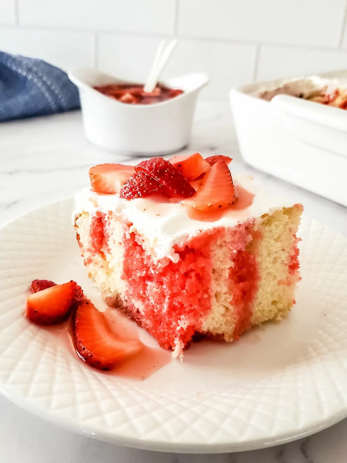 Slice of strawberry poke cake with fresh strawberries on top. In the background is a bowl of the strawberry sauce and the rest of the cake in a white dish.