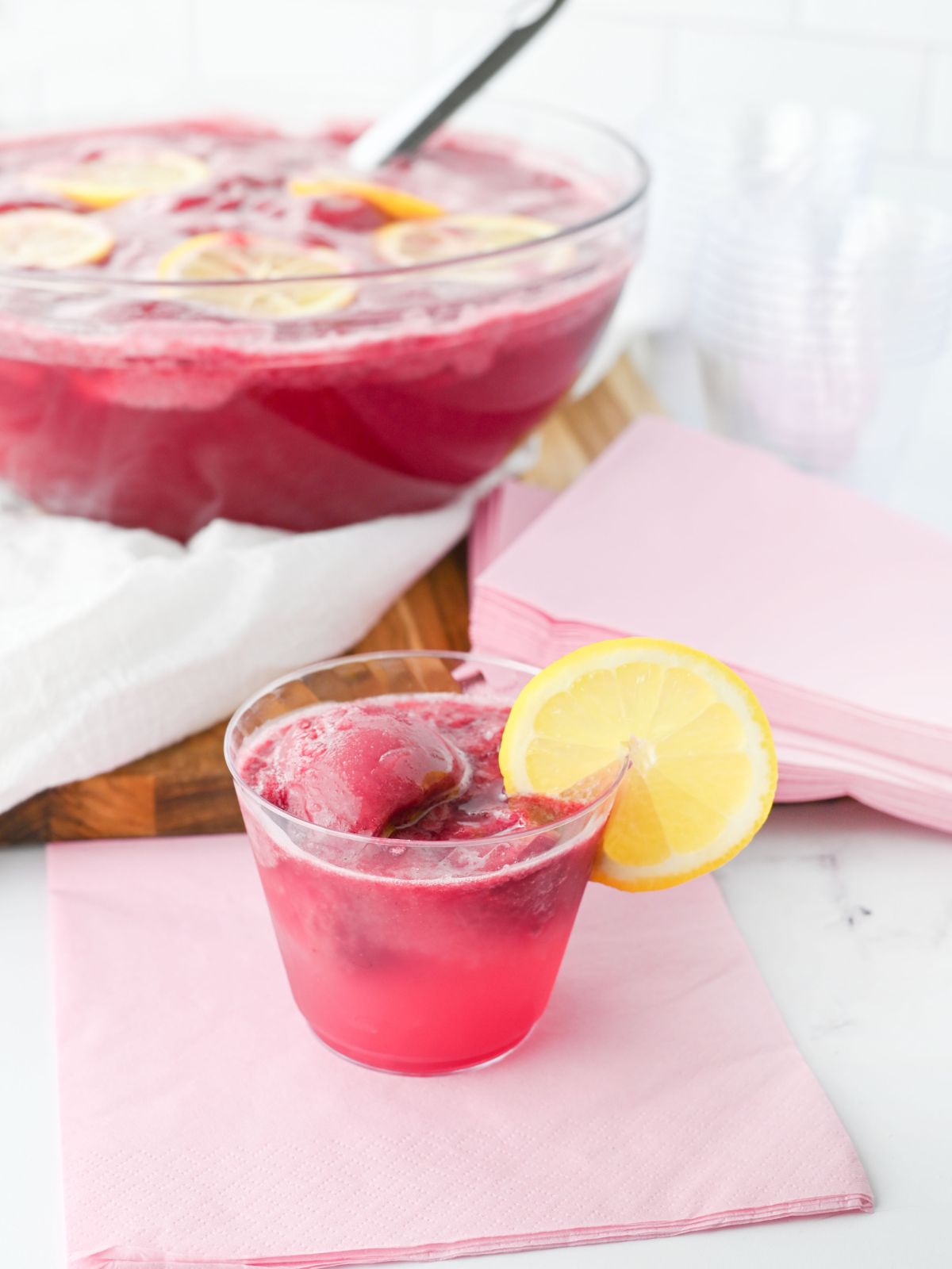 punch cup filled with raspberry punch garnished with a lemon wedge with the punch bowl in the background