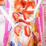 A close-up of a plastic bag filled with muffins topped with strawberry pie filling, whipped cream, and fresh strawberry slices.