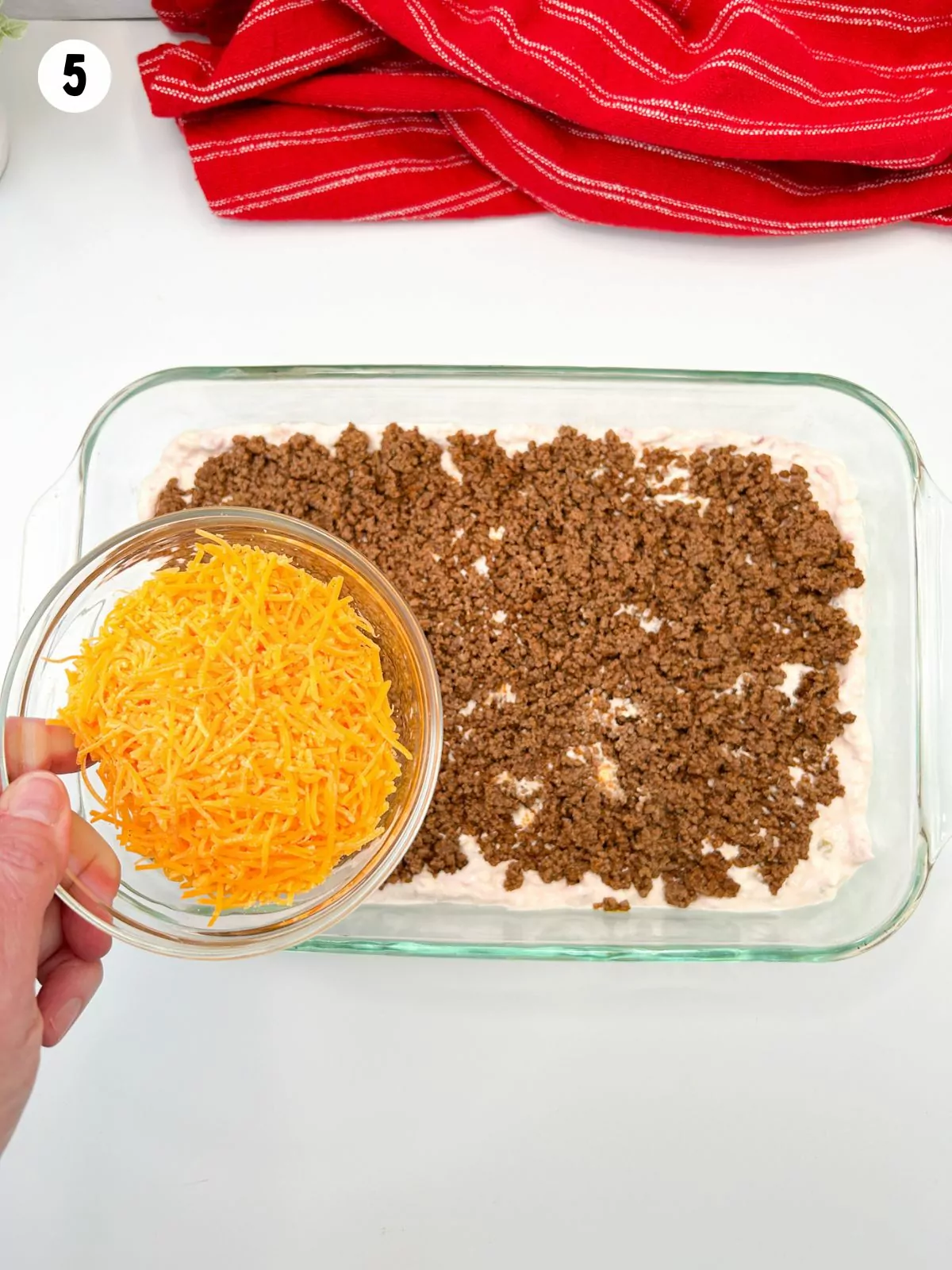 small bowl of cheddar cheese being held over taco dip with meat and cream cheese mixture.