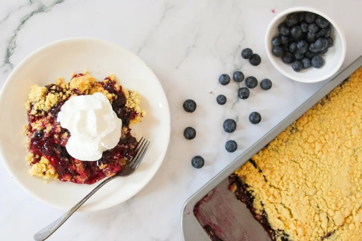 a plate of dump cake with blueberries and whipped cream next to a pan of baked cake.