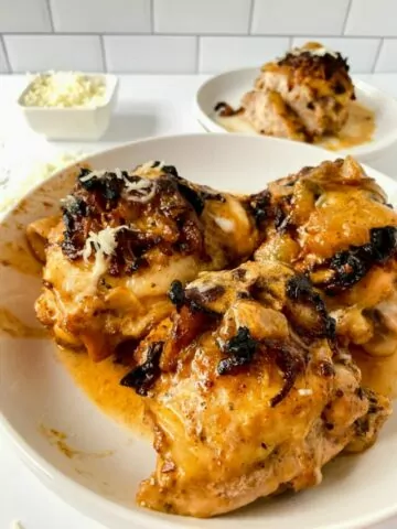 Baked chicken thighs with onions and cheese served on a white plate with a bowl of cheese and another plate of chicken in the background.