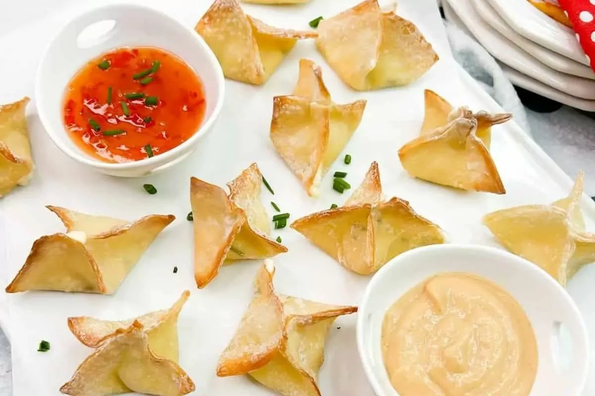 a plate of wonton wrappers filled with savory cream cheese with 2 small bowls of dipping sauces.