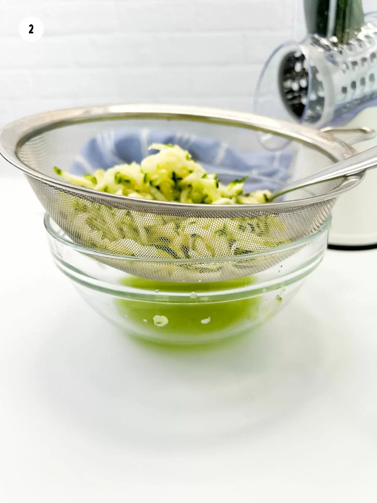 grated zucchini being drained in metal sieve over glass bowl filled with green juice.