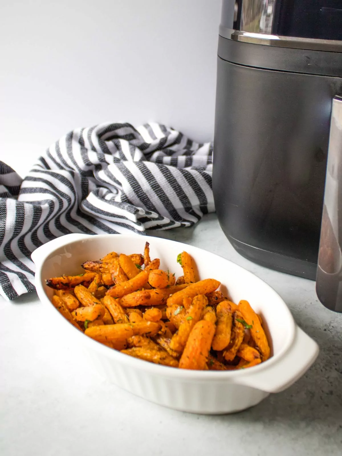 Baby carrots served in a white oval dish with an air fryer and striped kitchen towel in the background.